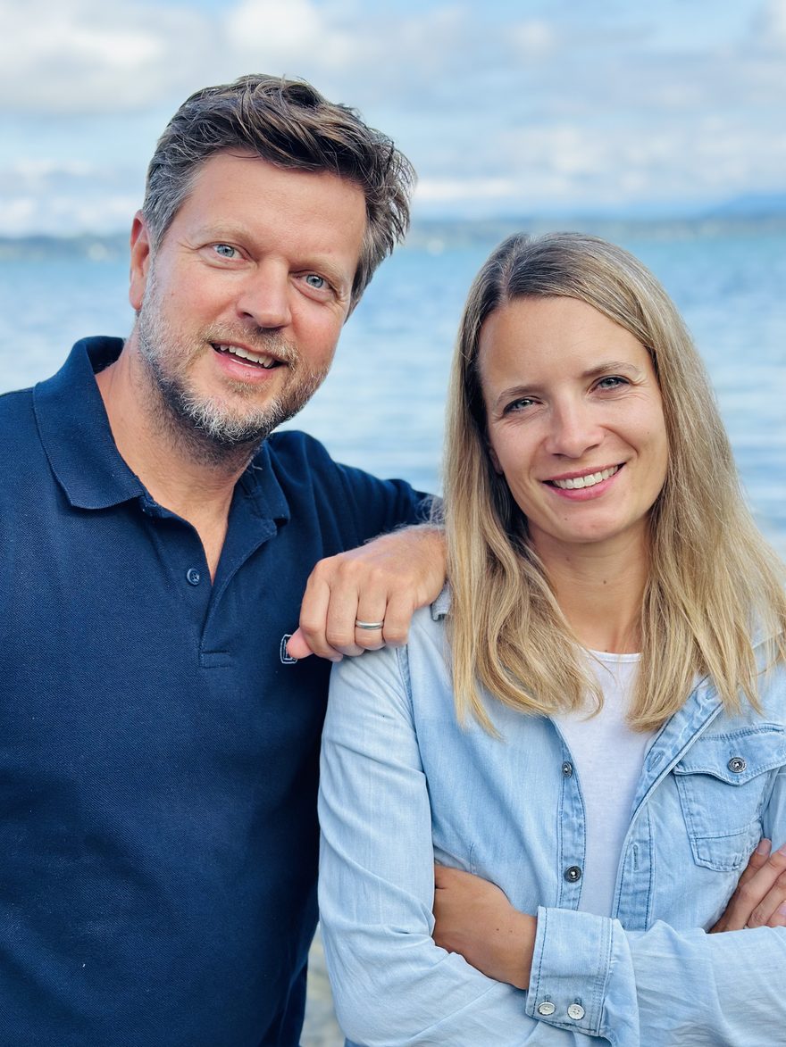 Melanie und Robert nebeneinander am Ufer des Starnberger Sees, beide lachen in die Kamera.
